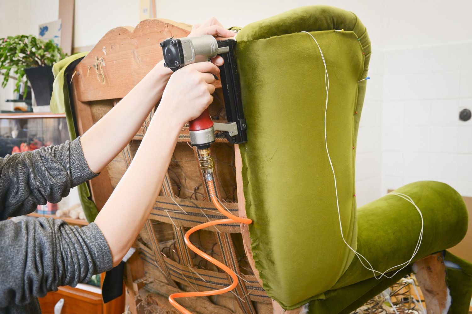A woman repairs a green chair with a drill in a cozy workshop, surrounded by tools and upholstery materials.