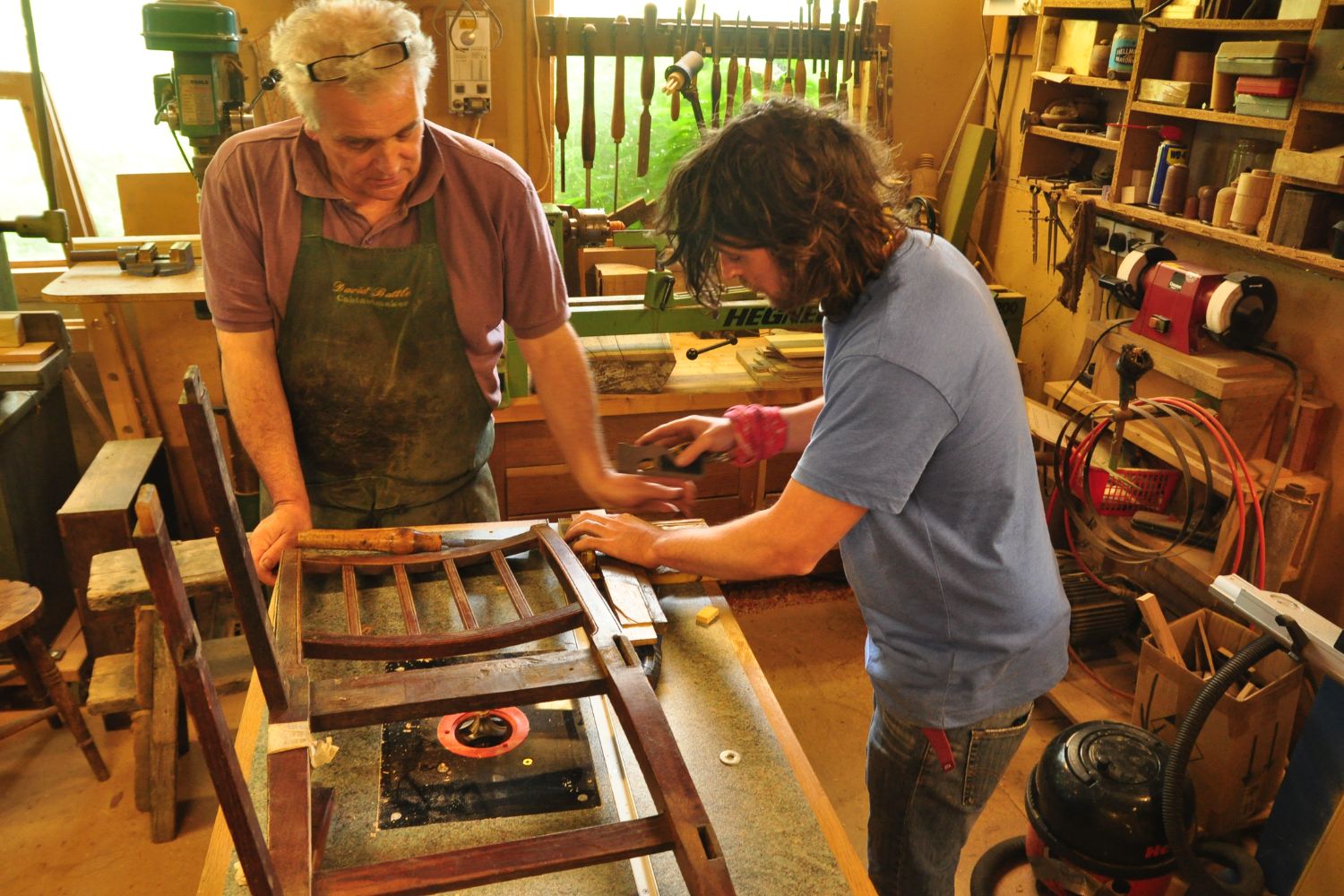 Two people work together on a wooden chair in a sunlit workshop, surrounded by tools and crafting supplies.