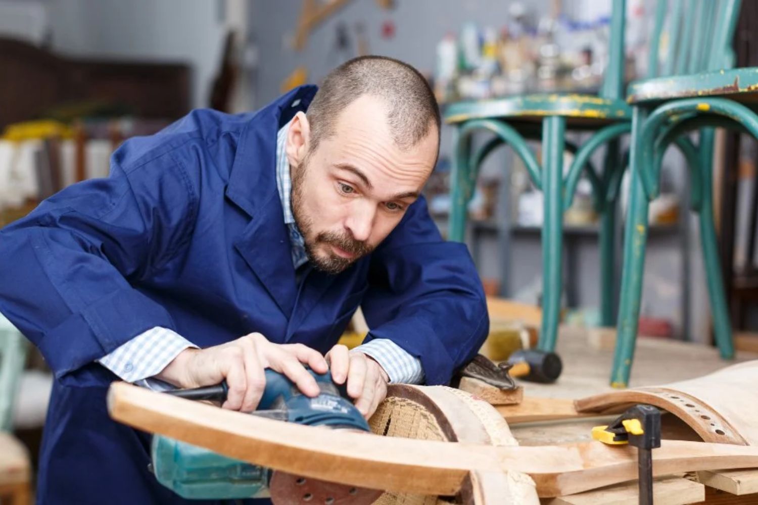 A craftsman in a blue work coat meticulously sands a wooden chair leg in a workshop, surrounded by tools and furniture.