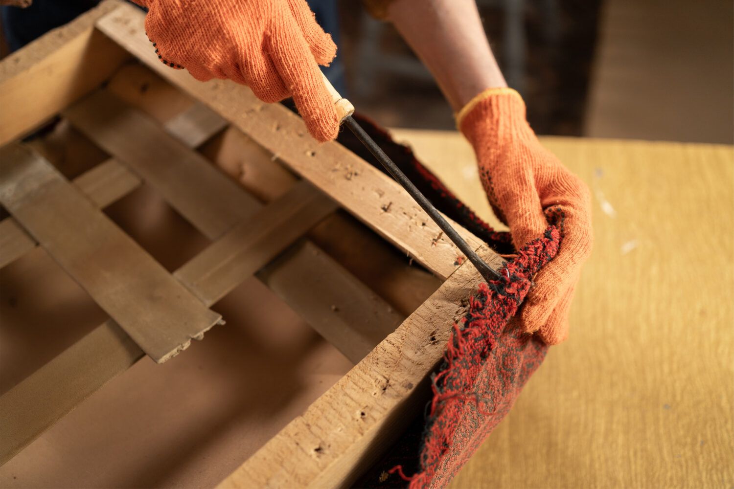 Person wearing orange gloves carefully removing fabric from a wooden furniture frame with a tool, surrounded by upholstery materials.