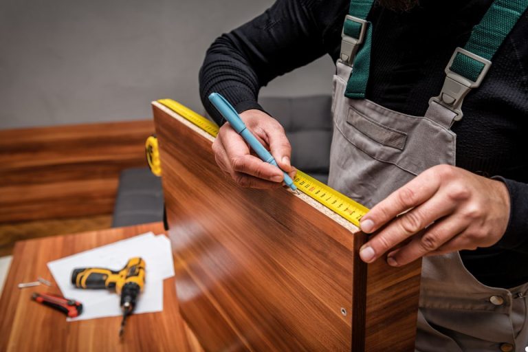 A man measures a piece of wood on a table using a ruler, focusing intently on the measurement.